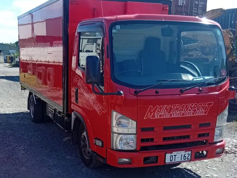 Red Main Transport Isuzu box truck parked in front of stacked shipping containers at the Suva depot