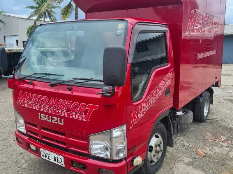 Red Main Transport Isuzu box truck, front three-quarter view at the Suva depot, palm trees behind